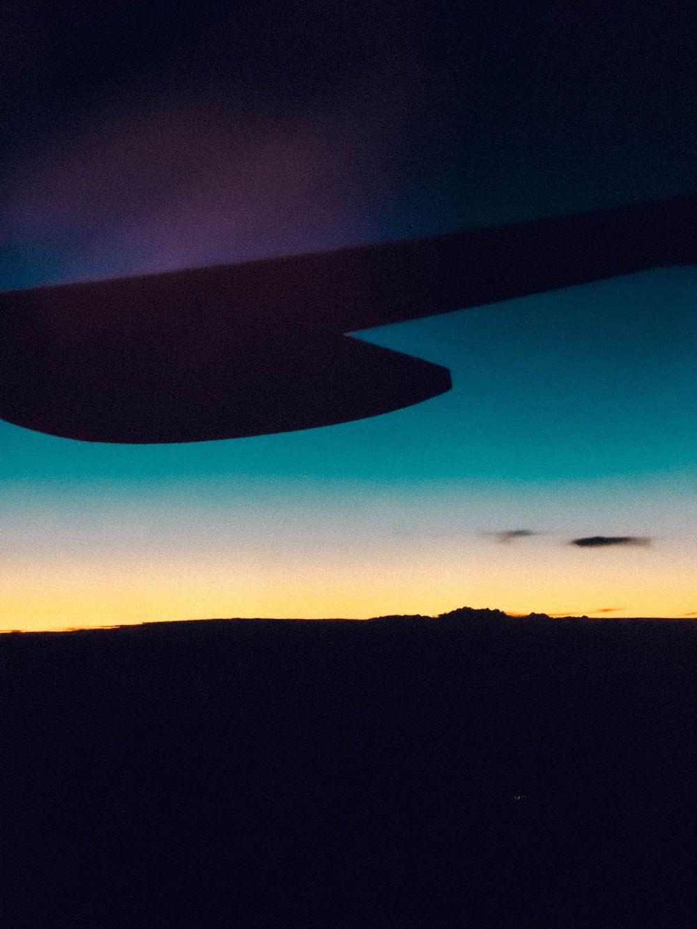 View from airplane window at sunset showing dark silhouette of wing against turquoise and orange gradient sky.