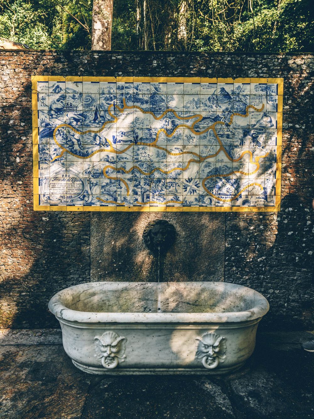 Vintage stone bathtub with lion head spouts against a weathered wall in dappled sunlight.