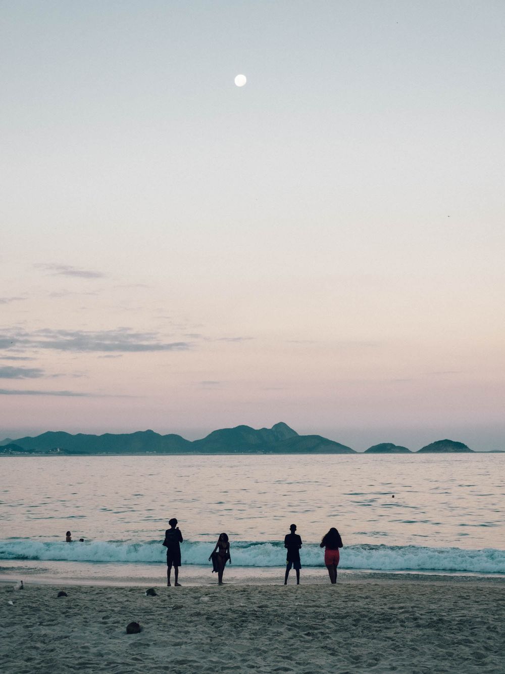Silhouettes of people walking along a scenic beach at sunset with mountains in the distance and calm ocean waters.