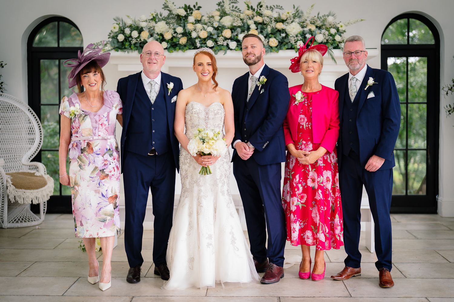Wedding group portrait in front of a bright white venue with floral arch decorations. Woodhill Hall.