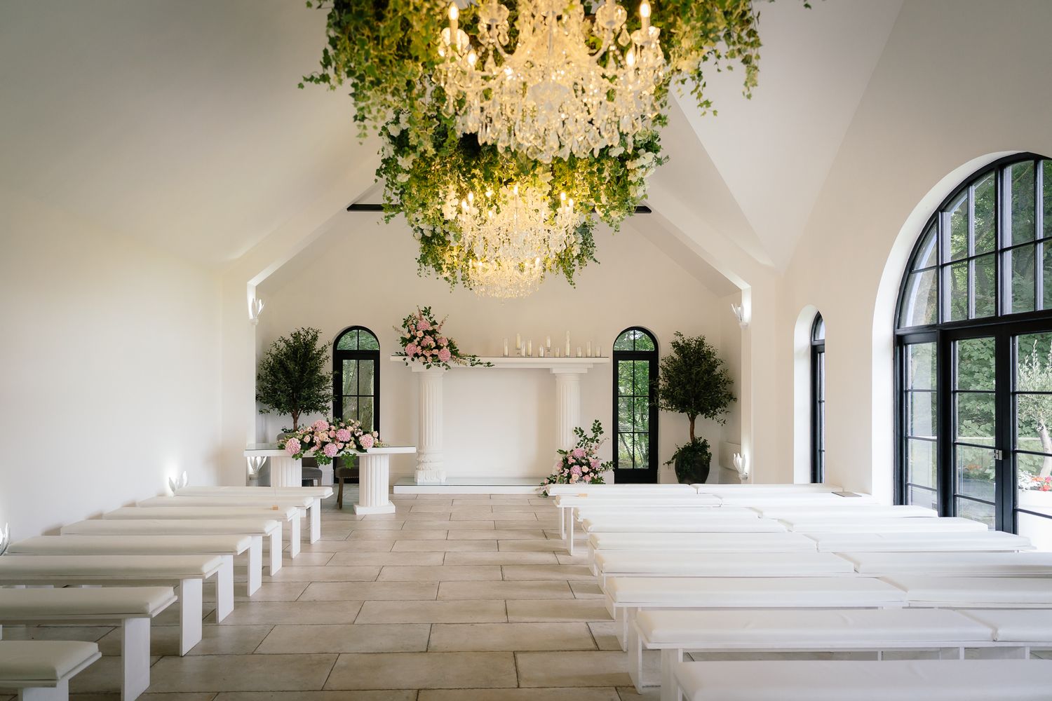 Modern white chapel interior with hanging floral chandelier and tiered seating for ceremonies. Woodhill Hall.