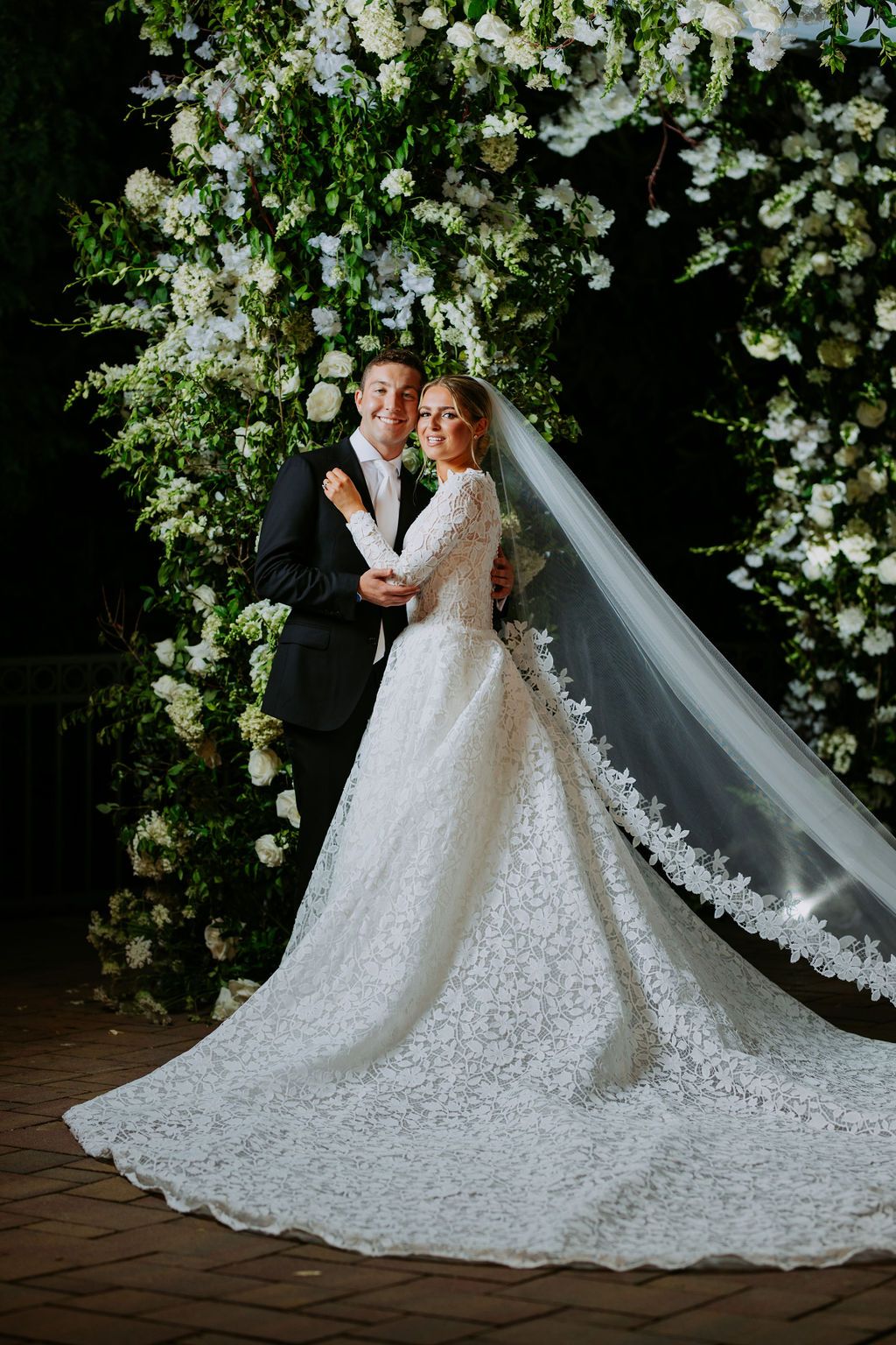 A flowing white wedding veil with delicate lace trim trails across a brick pathway in an outdoor garden setting.