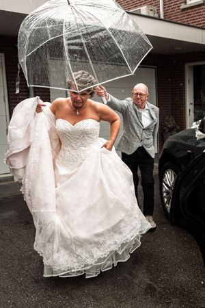 A bride in a white wedding dress holds a clear umbrella while walking through the rain on her special day.