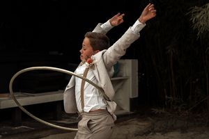 A person in a white shirt and vest performs with a hula hoop on stage against a dark background.