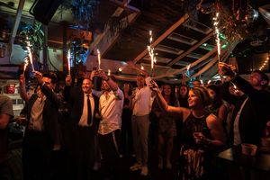 A lively crowd dances and celebrates with raised arms in a dimly lit indoor venue during a party or event.