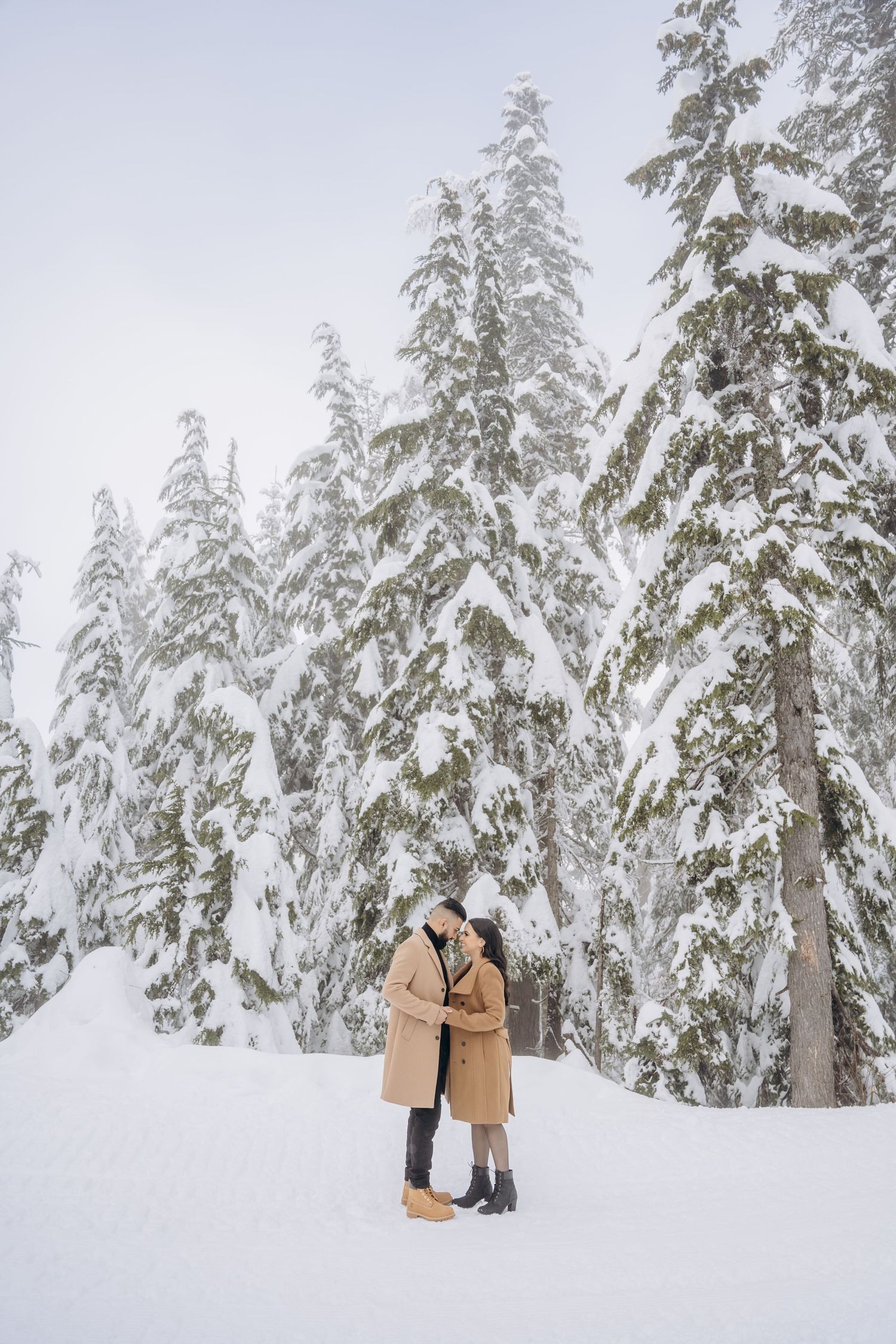 A couple embraces in a snowy winter forest with snow-covered evergreen trees creating a romantic backdrop.