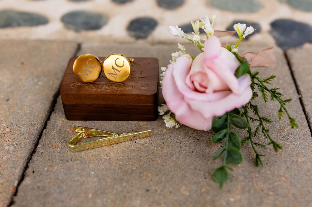 Single pink rose with gold wedding rings in wooden box on stone surface.