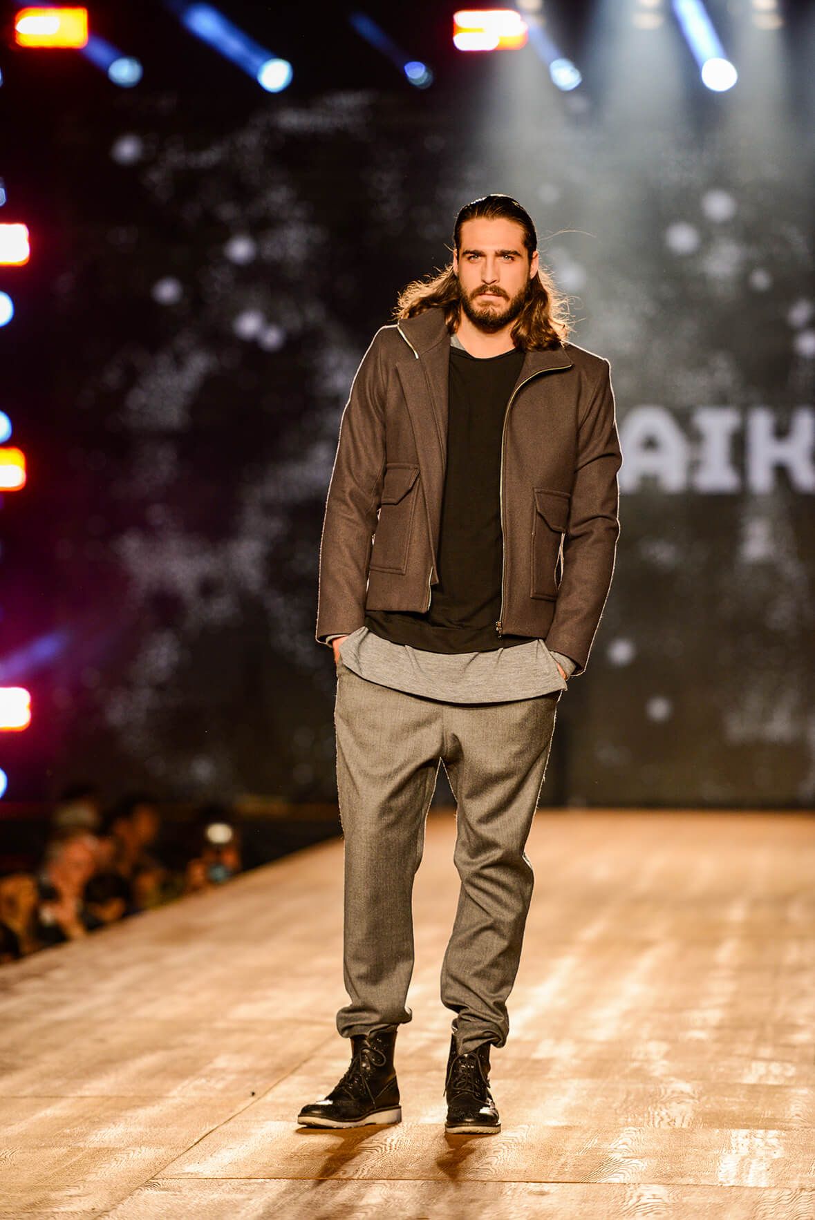 Model walks runway in brown jacket and gray joggers during fashion show against dark illuminated backdrop. Montreal, Canada, Wedding Photos