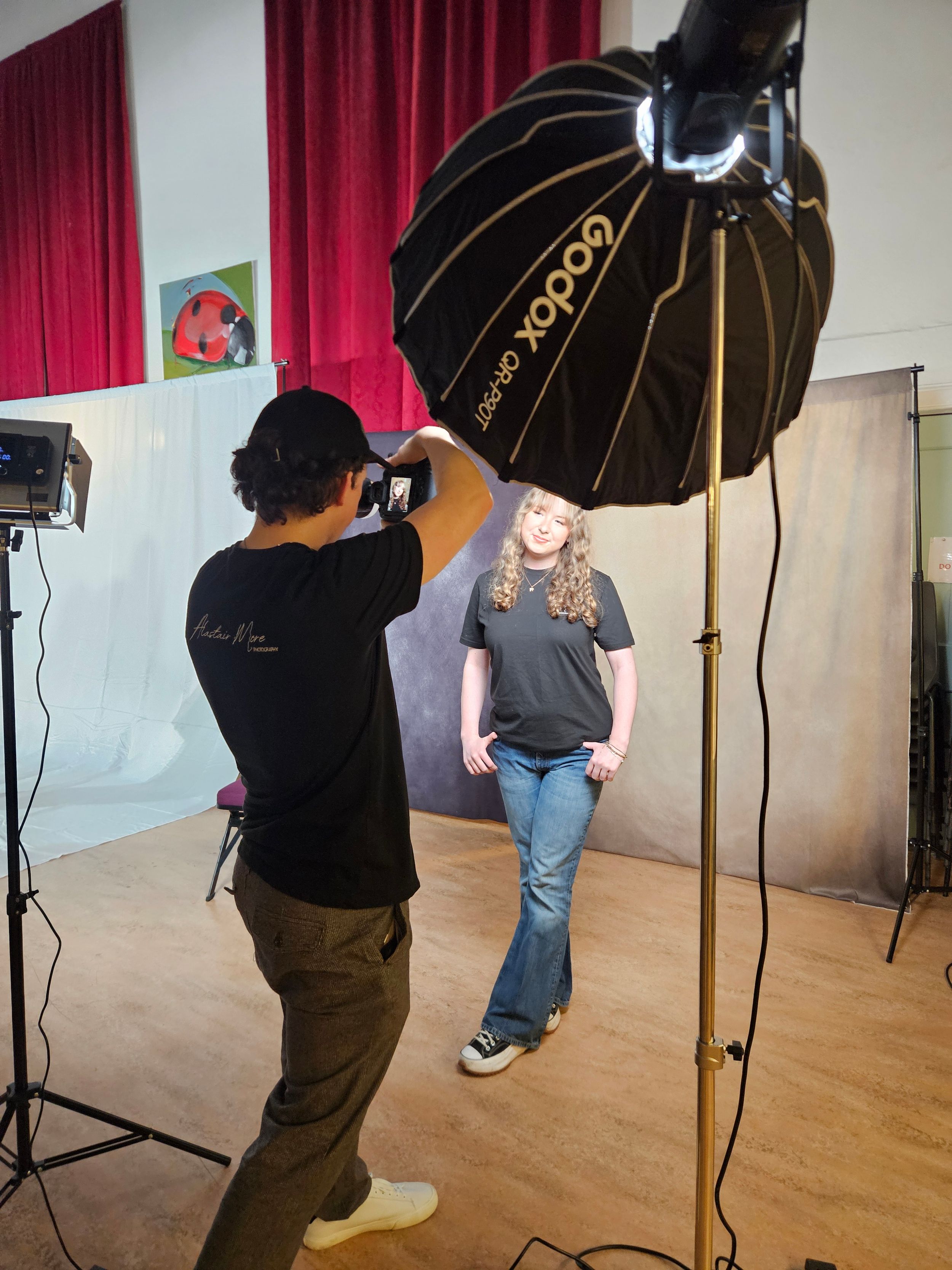 Professional photo shoot setup in studio with large black umbrella light modifier and photographer capturing model against red curtain.