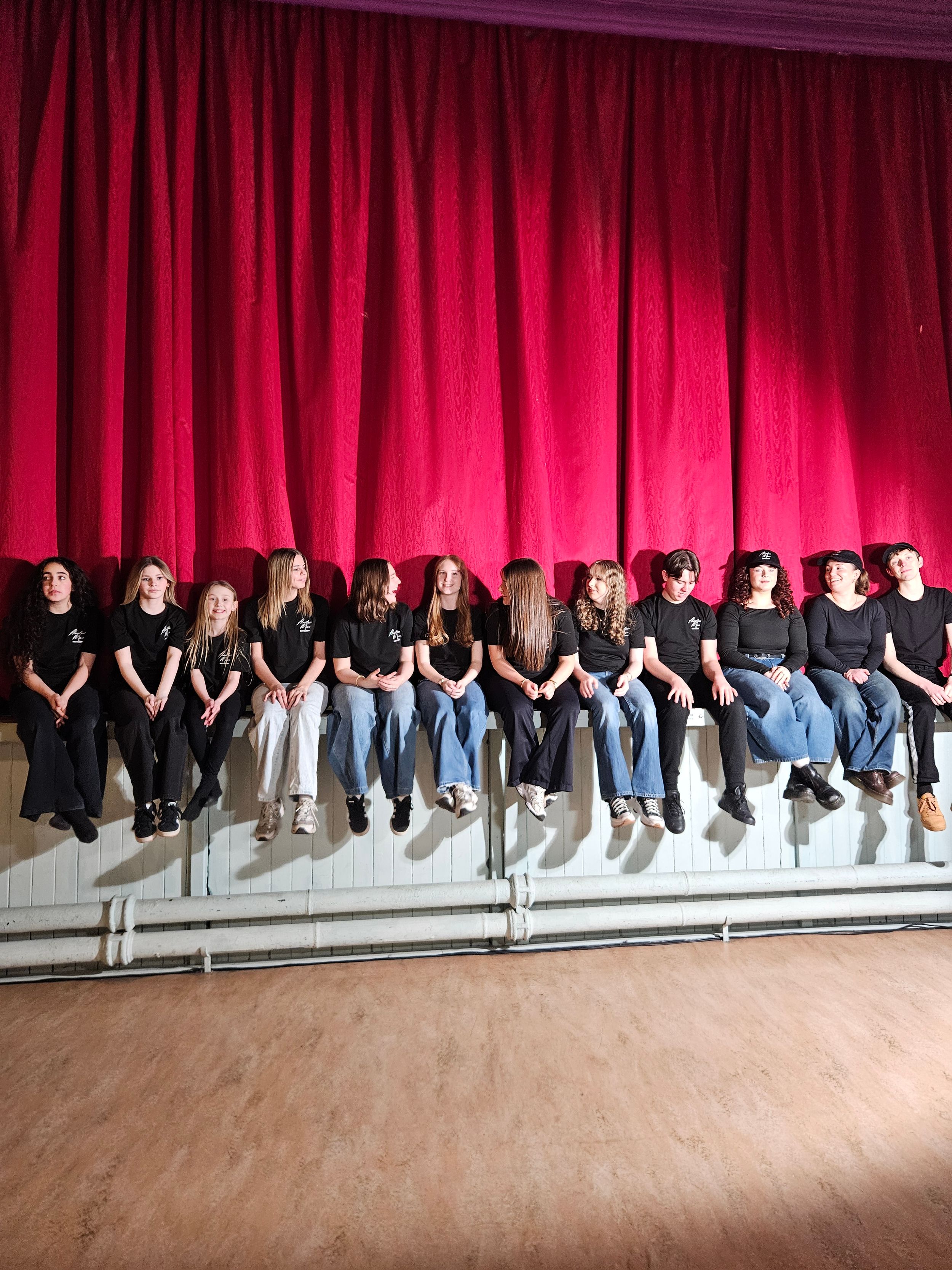 A row of performers in black and blue outfits sit along the edge of a stage with a red curtain backdrop.