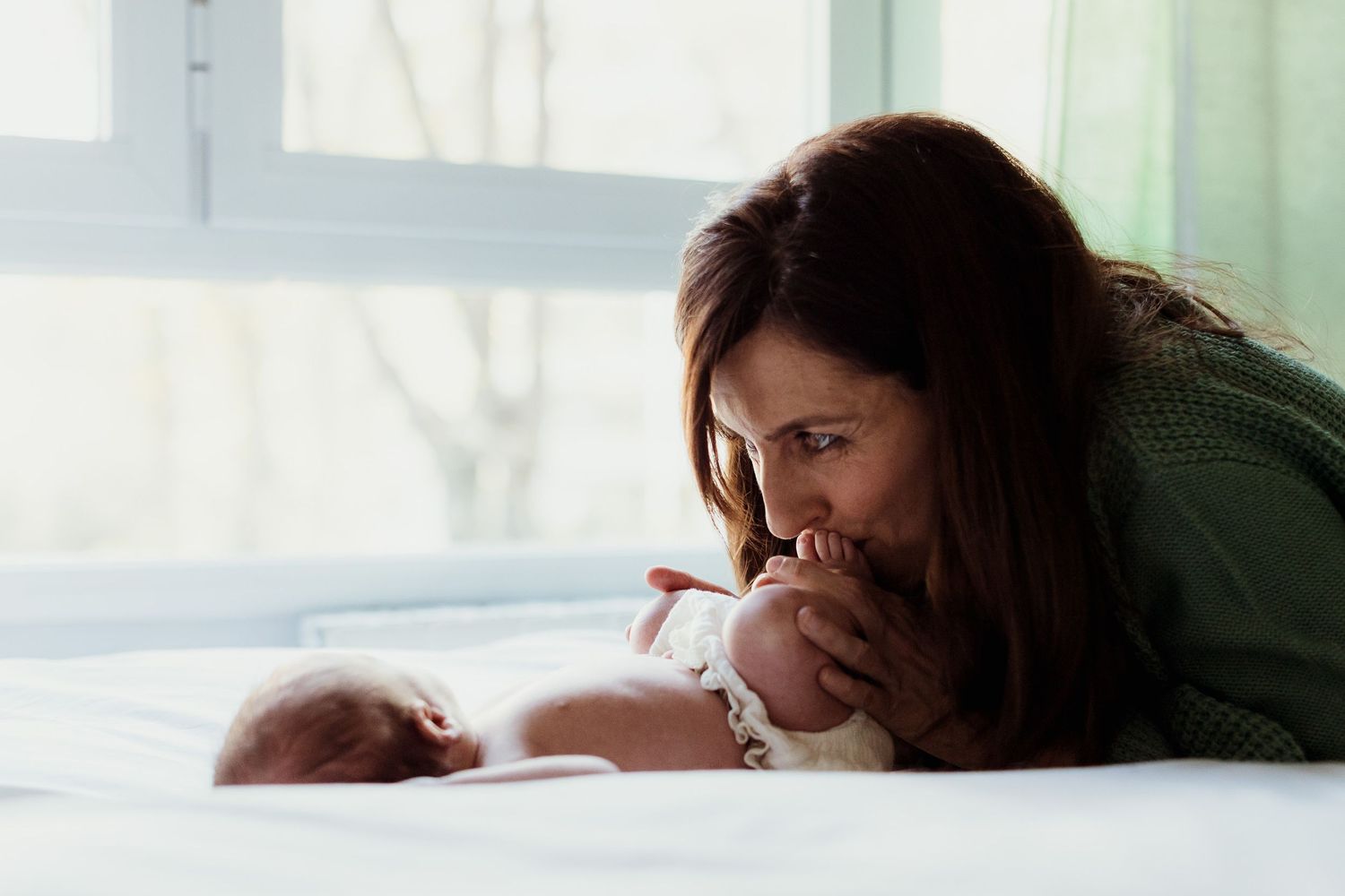 A tender moment shared between parent and baby lying together on a white bed near a bright window.