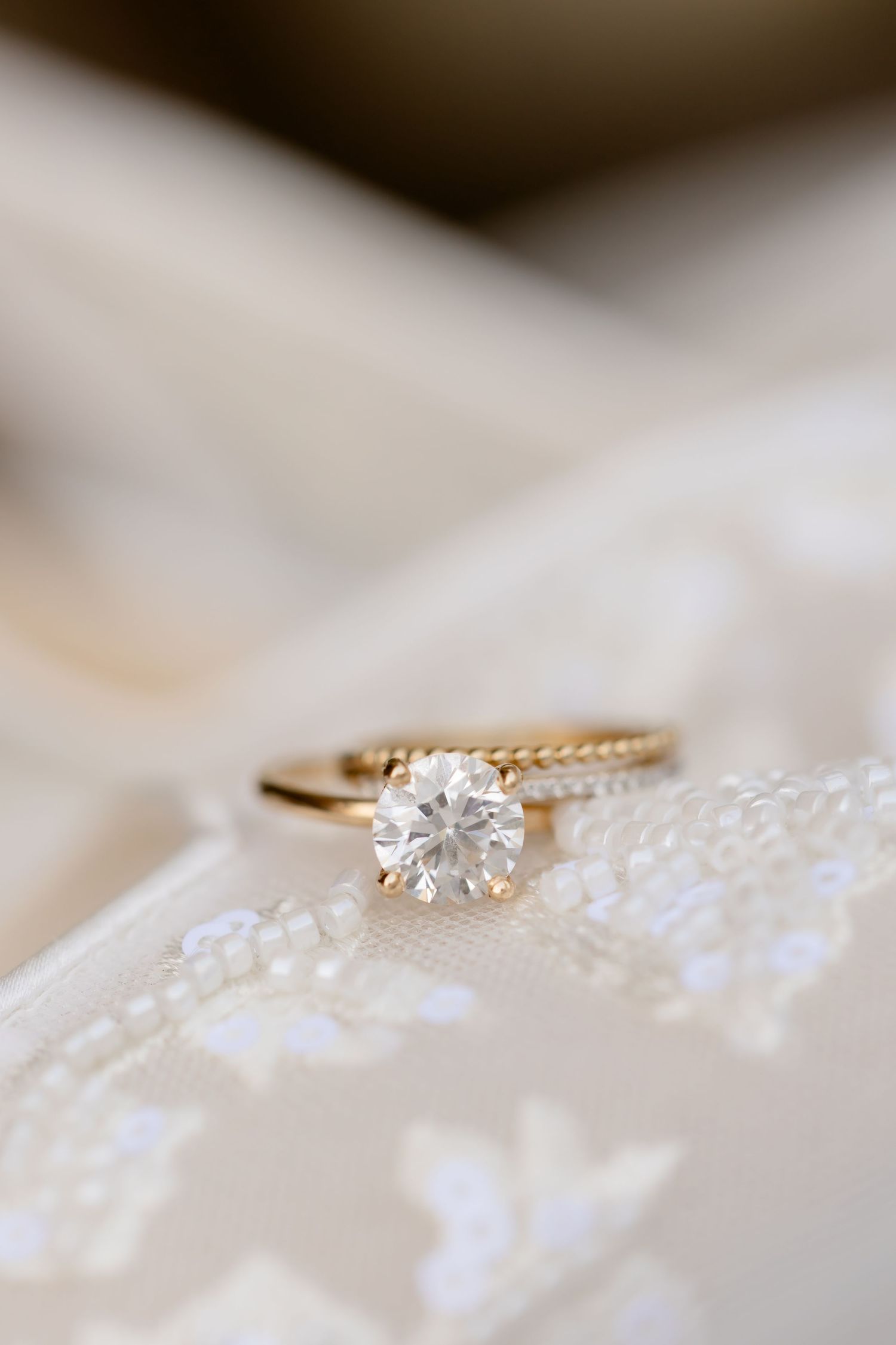 A delicate diamond engagement ring rests on white lace fabric with soft bokeh lighting effects in the background.