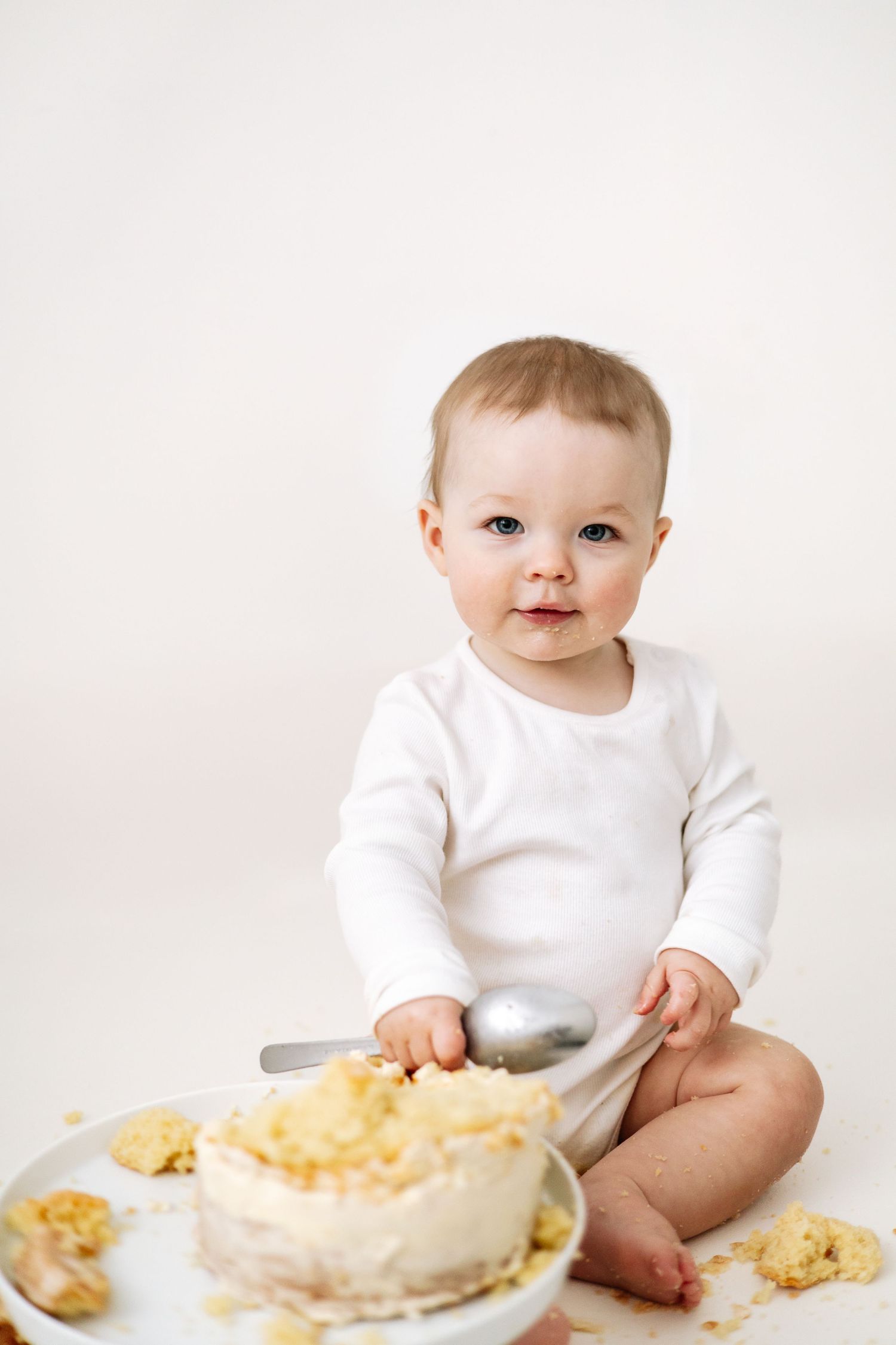 Baby in white onesie sits on floor with messy cake, during Birthday cake smash shoot based in Nottingham