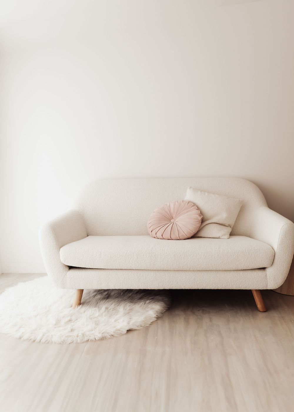 Minimalist cream colored sofa with pink round pillow and white fluffy rug on light wooden floor against white wall.