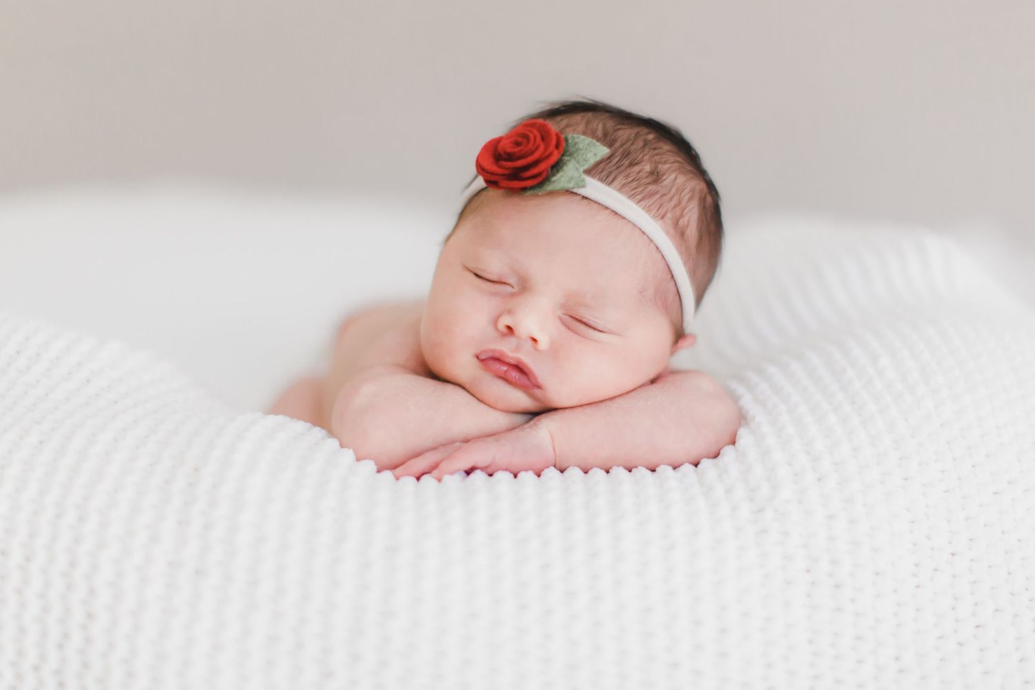 A sleeping newborn wearing a red rose headband on white textured fabric.
