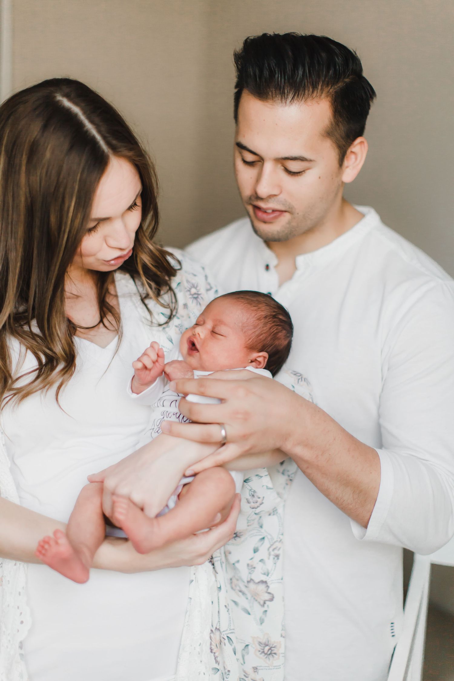 Parents in white clothing share intimate bonding moments with their newborn baby in a light-filled room.