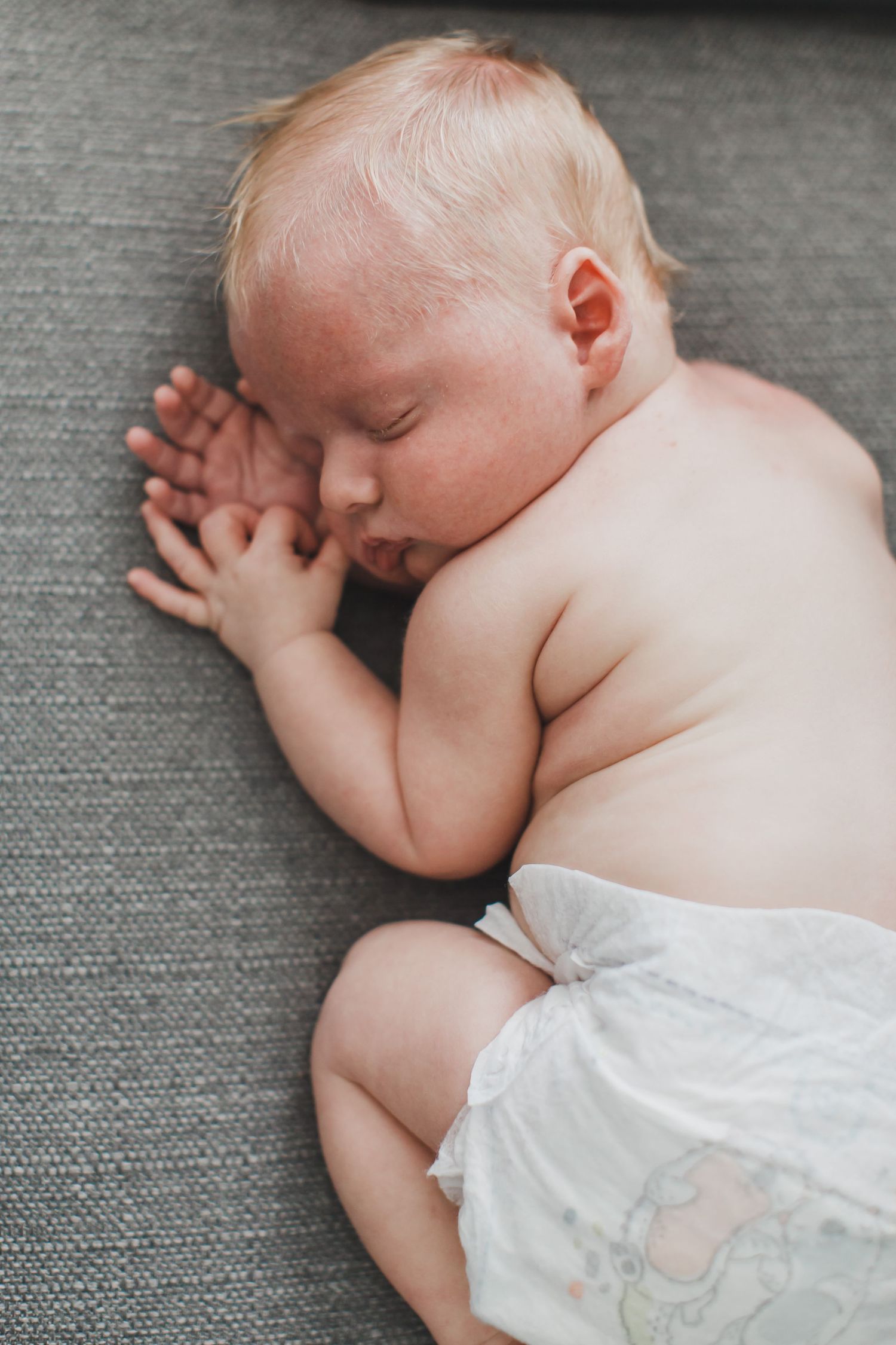 Sleeping newborn baby in diaper rests peacefully on grey textured fabric.