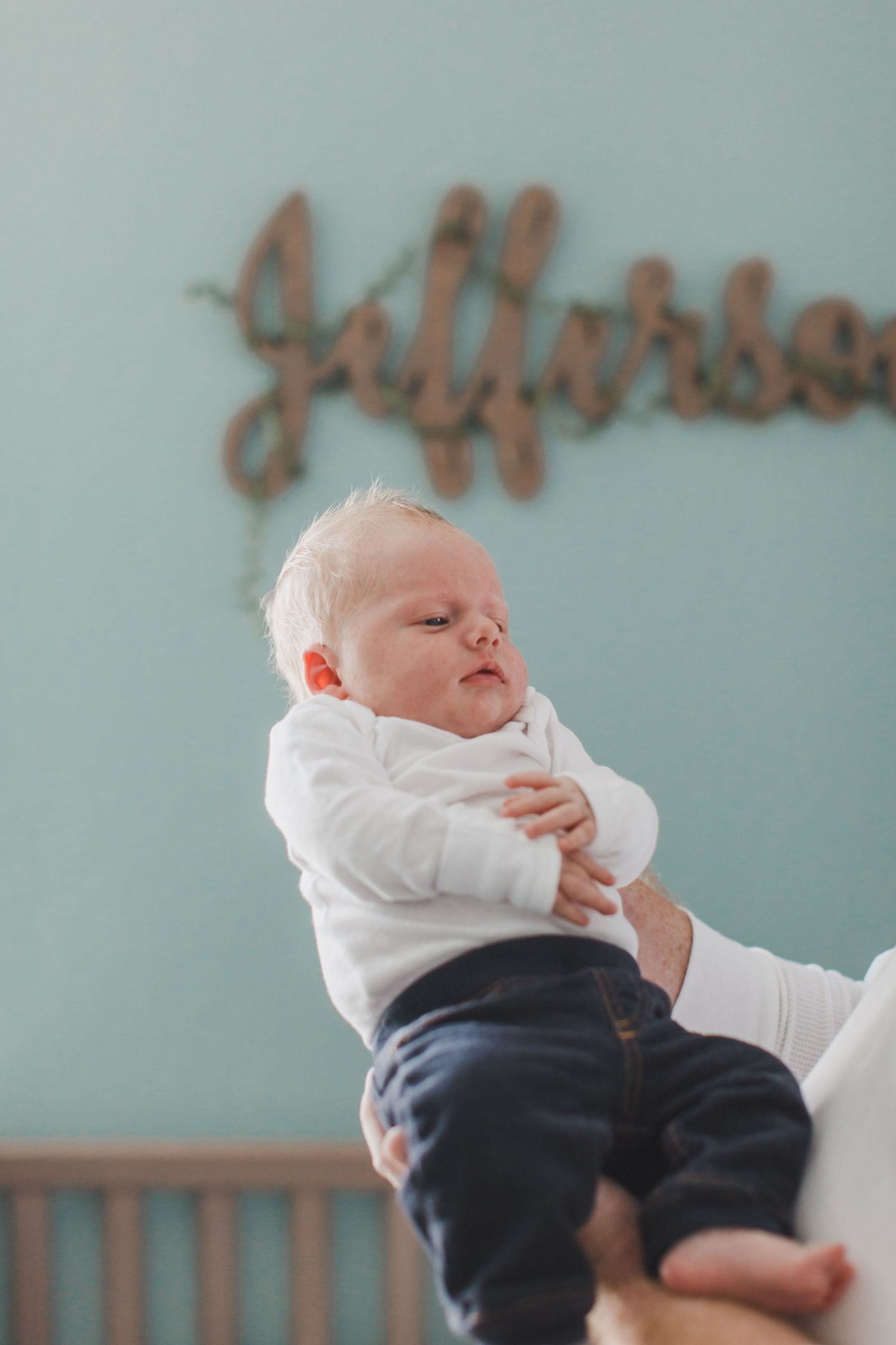 A newborn baby in white clothing and navy pants is held up against a soft blue wall.