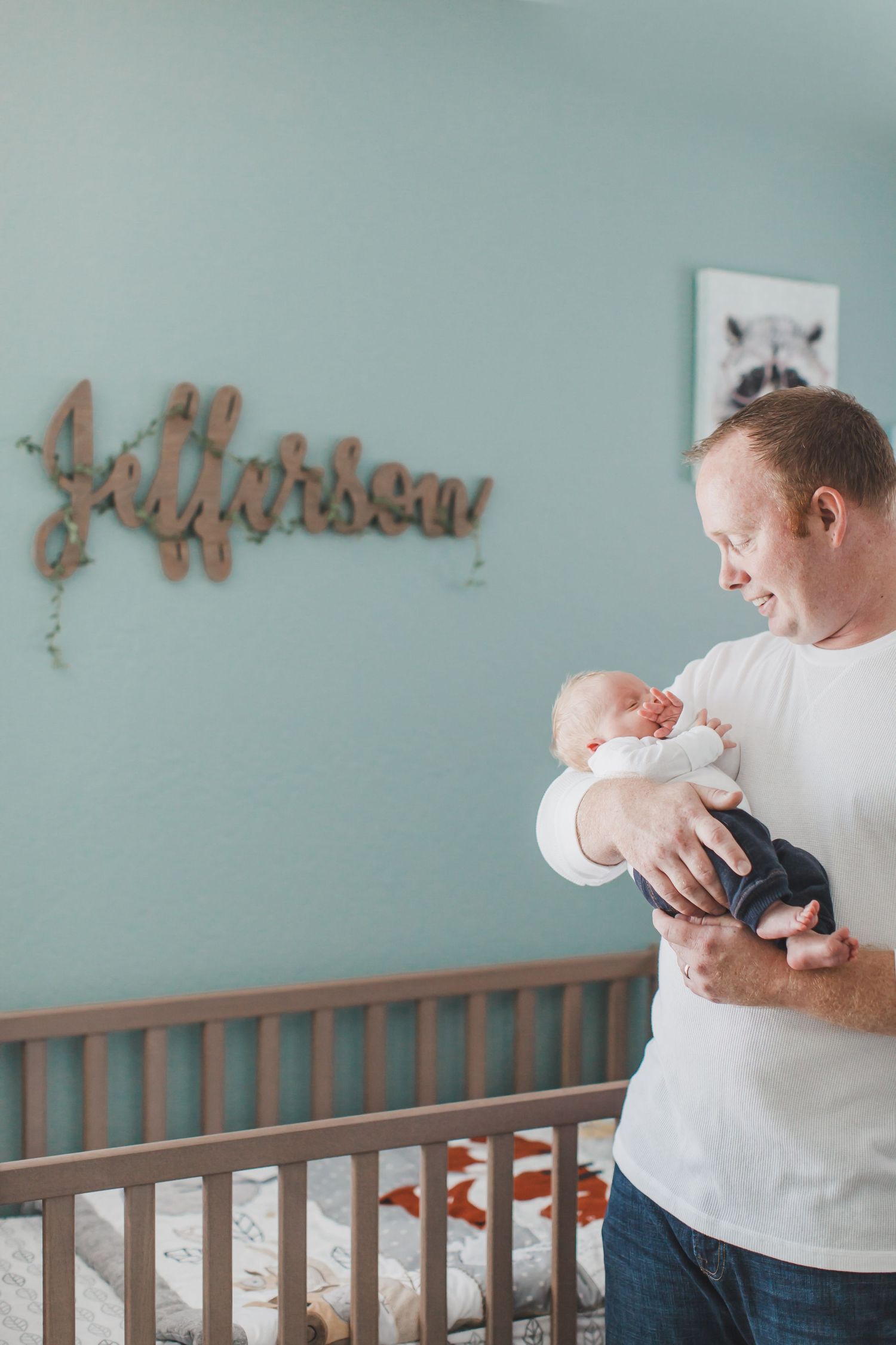 A parent in white holds a baby near a wooden crib with decorative wall art in the background.