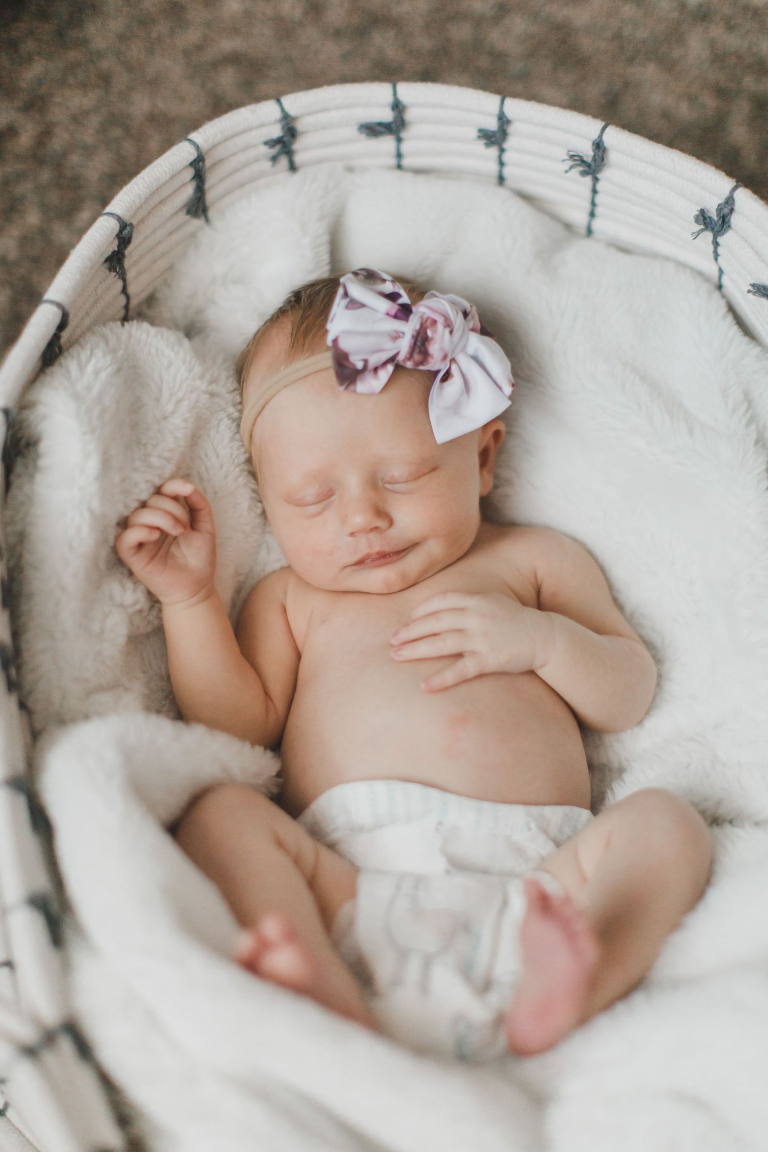 Sleeping newborn wearing purple bow on white cushioned basket.