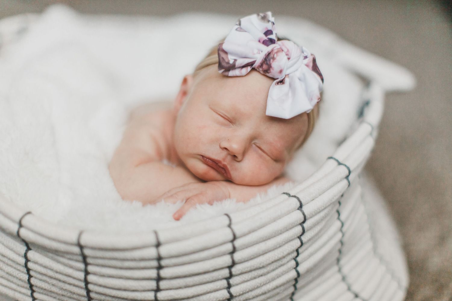 Sleeping newborn with bow accessory in cozy white basket setting.