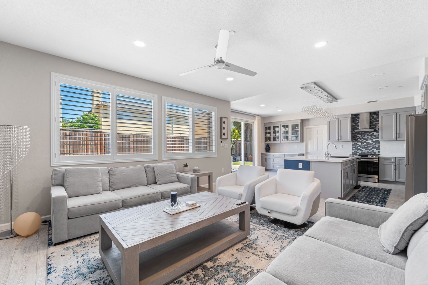 Modern open concept living room with grey sofas and connected kitchen featuring white cabinets and stainless appliances.
