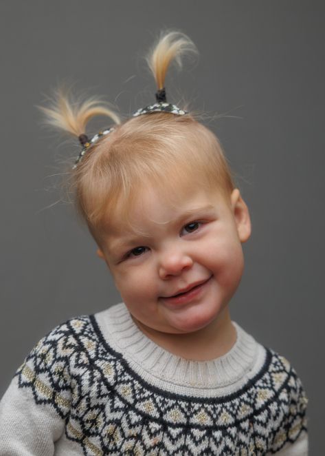 Small child wearing a Nordic style sweater with feathered hair clips smiling against a gray background.