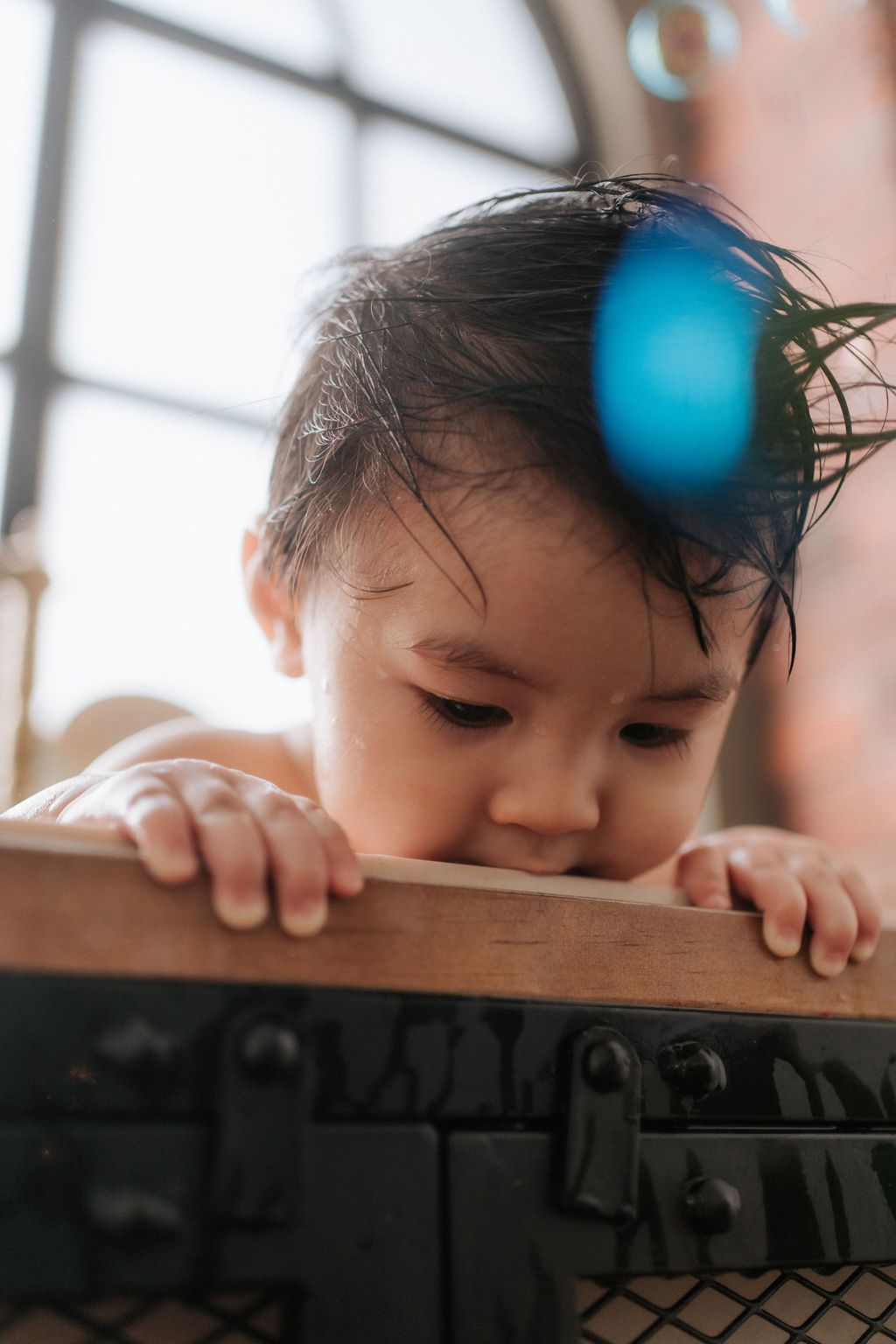 A curious child peeks over a wooden surface while looking down with soft window light illuminating their face.