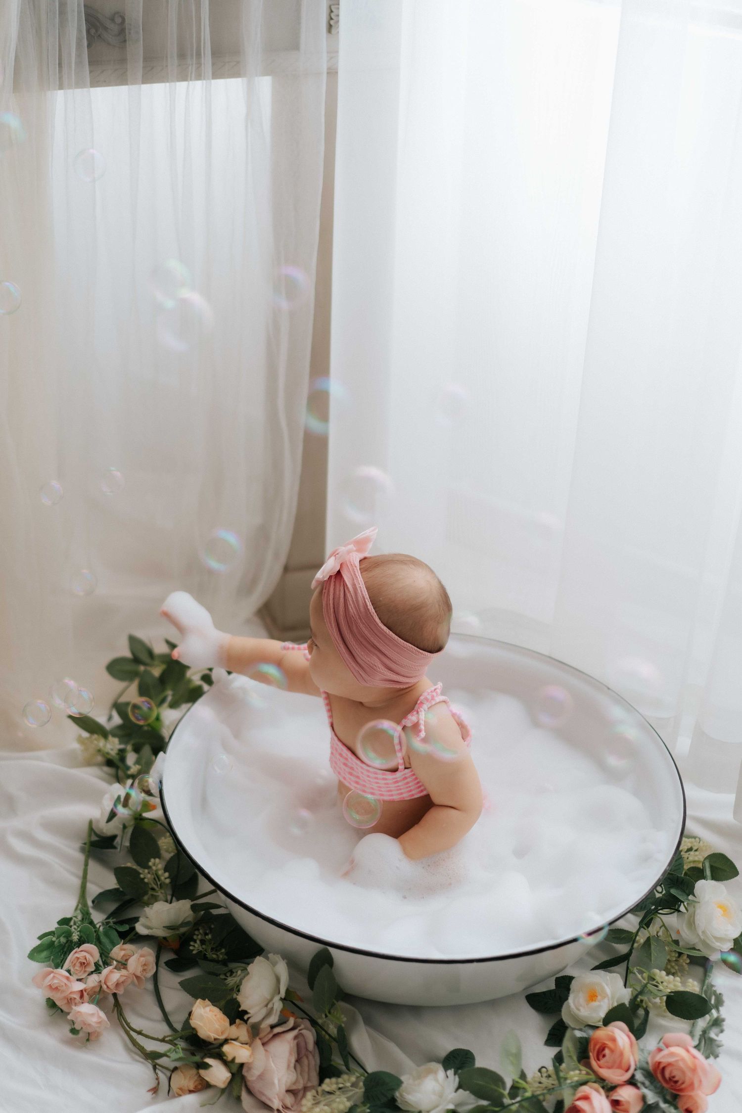 Baby in white outfit plays with colorful pastel balloons and smash cake during first birthday photoshoot.