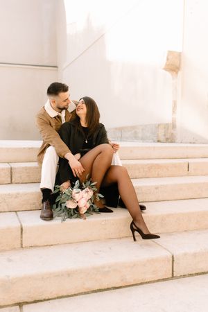 Couple romantique assis sur des marches blanches avec bouquet de fleurs rose dans un look élégant