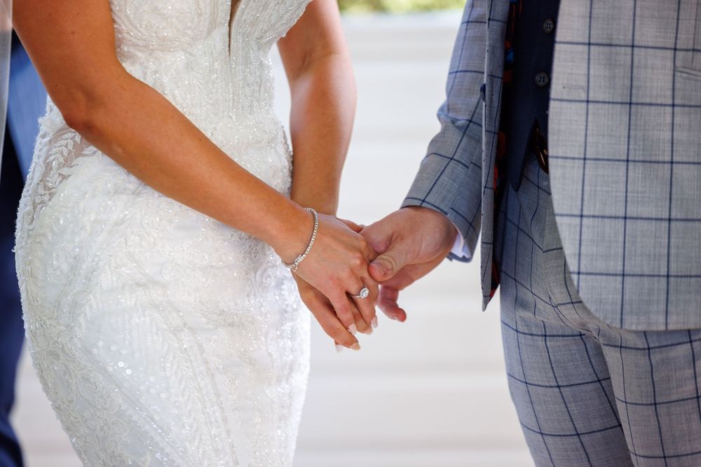 Bride and groom holding hands, wedding rings visible, bride in lace dress and groom in blue plaid suit.