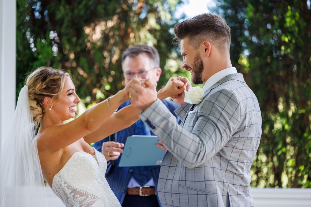 Bride placing wedding ring on grooms finger during outdoor ceremony with officiant holding blue book.