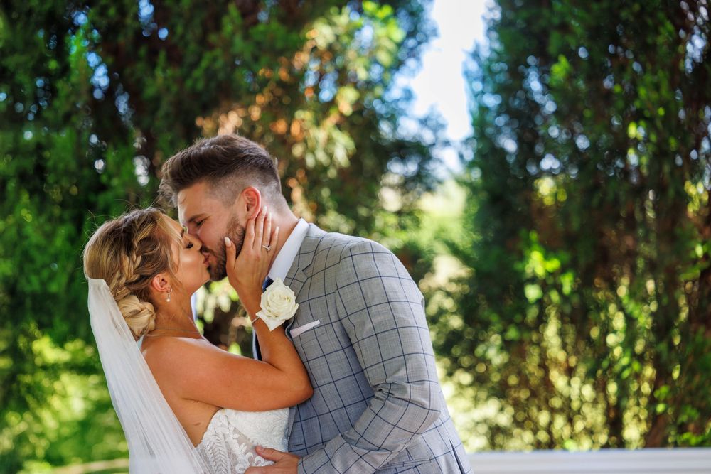 Bride and groom share first kiss surrounded by lush green trees on their wedding day.
