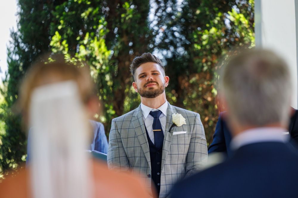 Groom in plaid suit watching bride walk down the aisle at outdoor wedding ceremony.