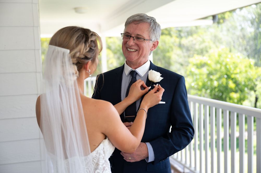 Bride in lace gown pinning white boutonniere on fathers navy suit lapel before ceremony.