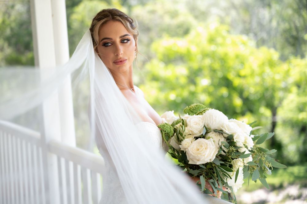 Bride in white wedding dress and veil holds bouquet of white roses on a sunlit porch.