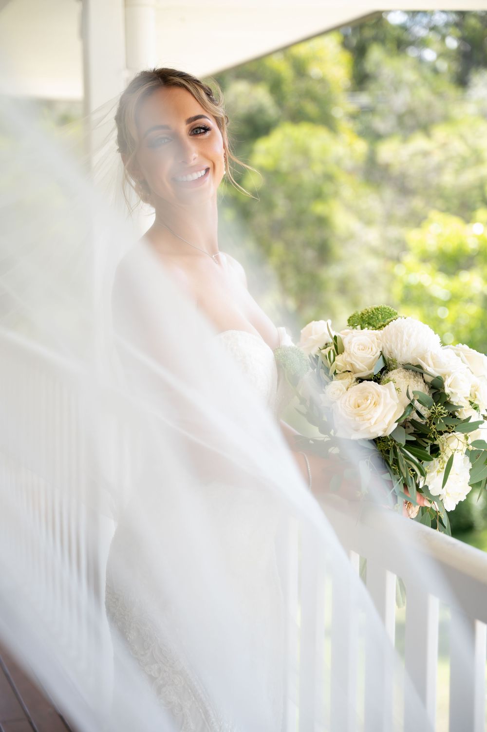 Smiling bride in flowing white veil holds white rose bouquet on a bright outdoor porch.