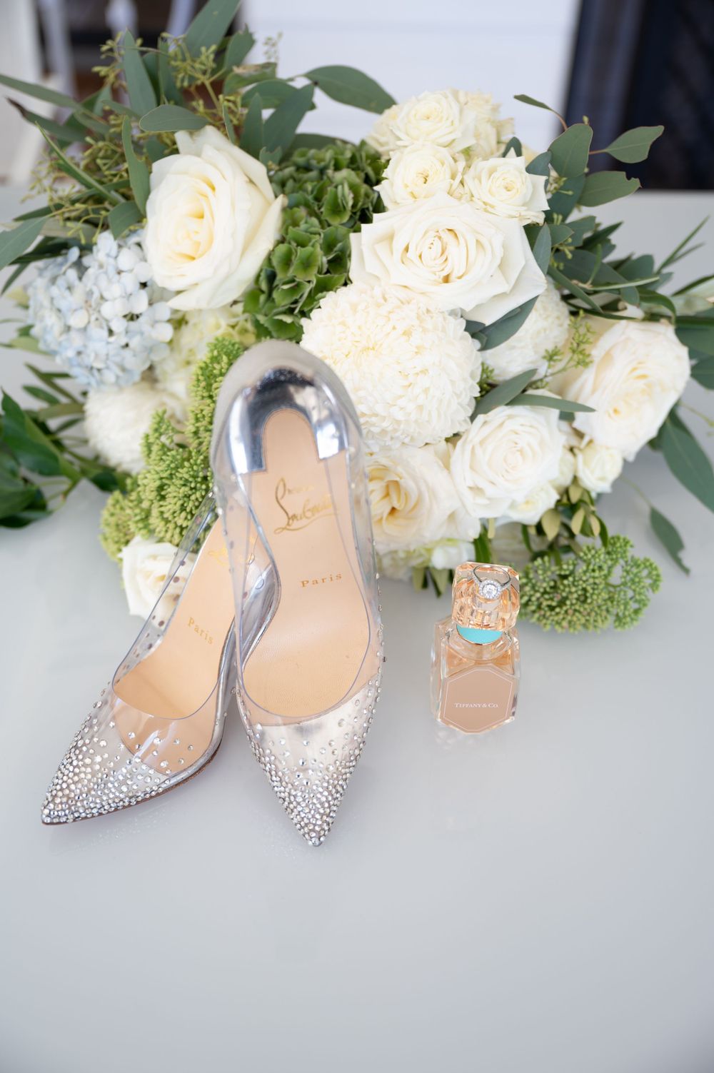 Crystal embellished Christian Louboutin bridal heels beside white rose and greenery wedding bouquet.