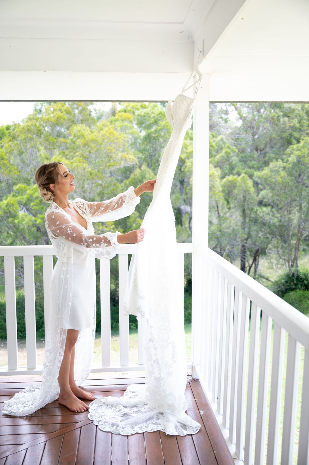 Bride in lace robe holding flowing wedding dress on white timber balcony surrounded by greenery.