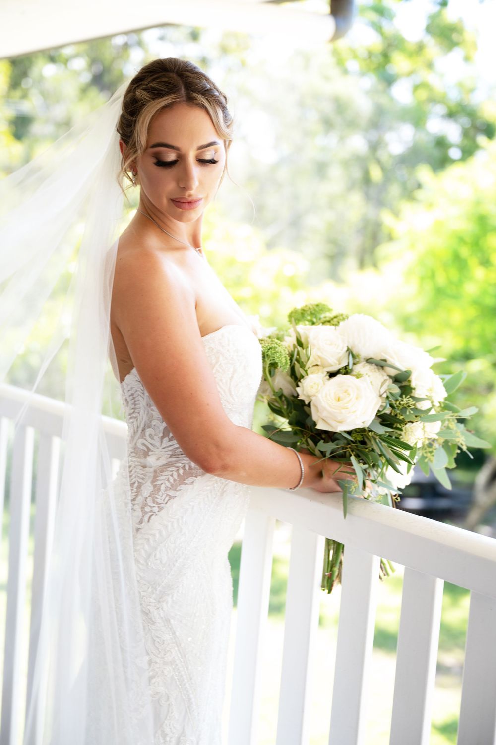 Beautiful bride in lace wedding gown holds white rose bouquet while standing on a white railing balcony outdoors.
