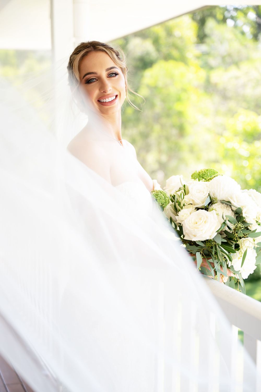 Radiant bride in flowing white veil laughs joyfully while holding a white floral bouquet on a sunlit balcony.