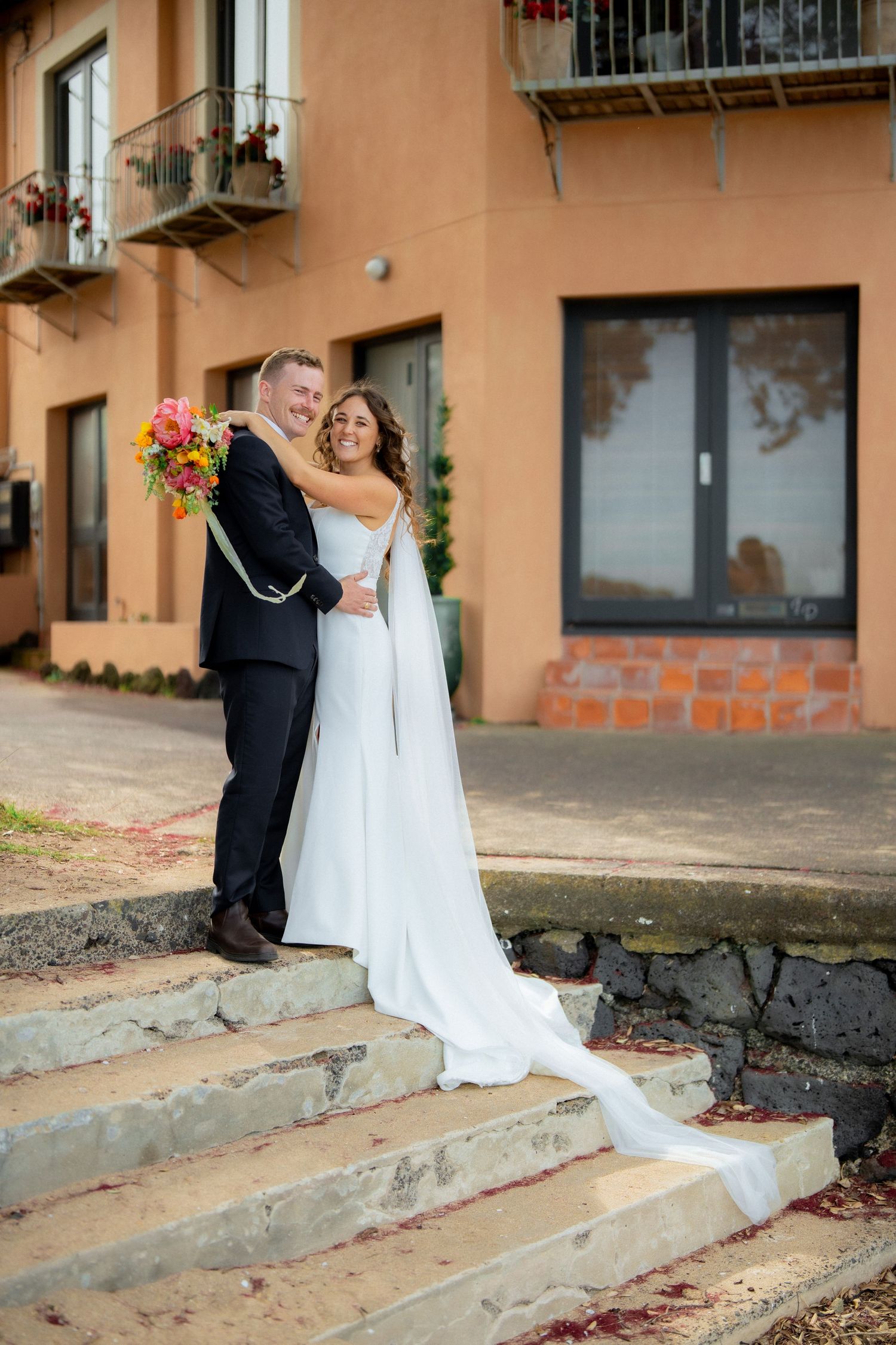 A couple in wedding attire embrace on stone steps outside a peach-colored building with trailing white dress and colorful bouquet.
