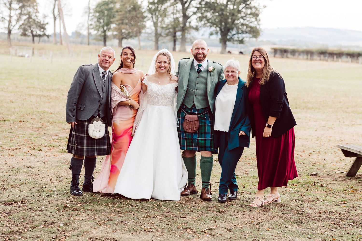 Wedding party members in green dresses and traditional Scottish kilts pose together outdoors on autumn day.