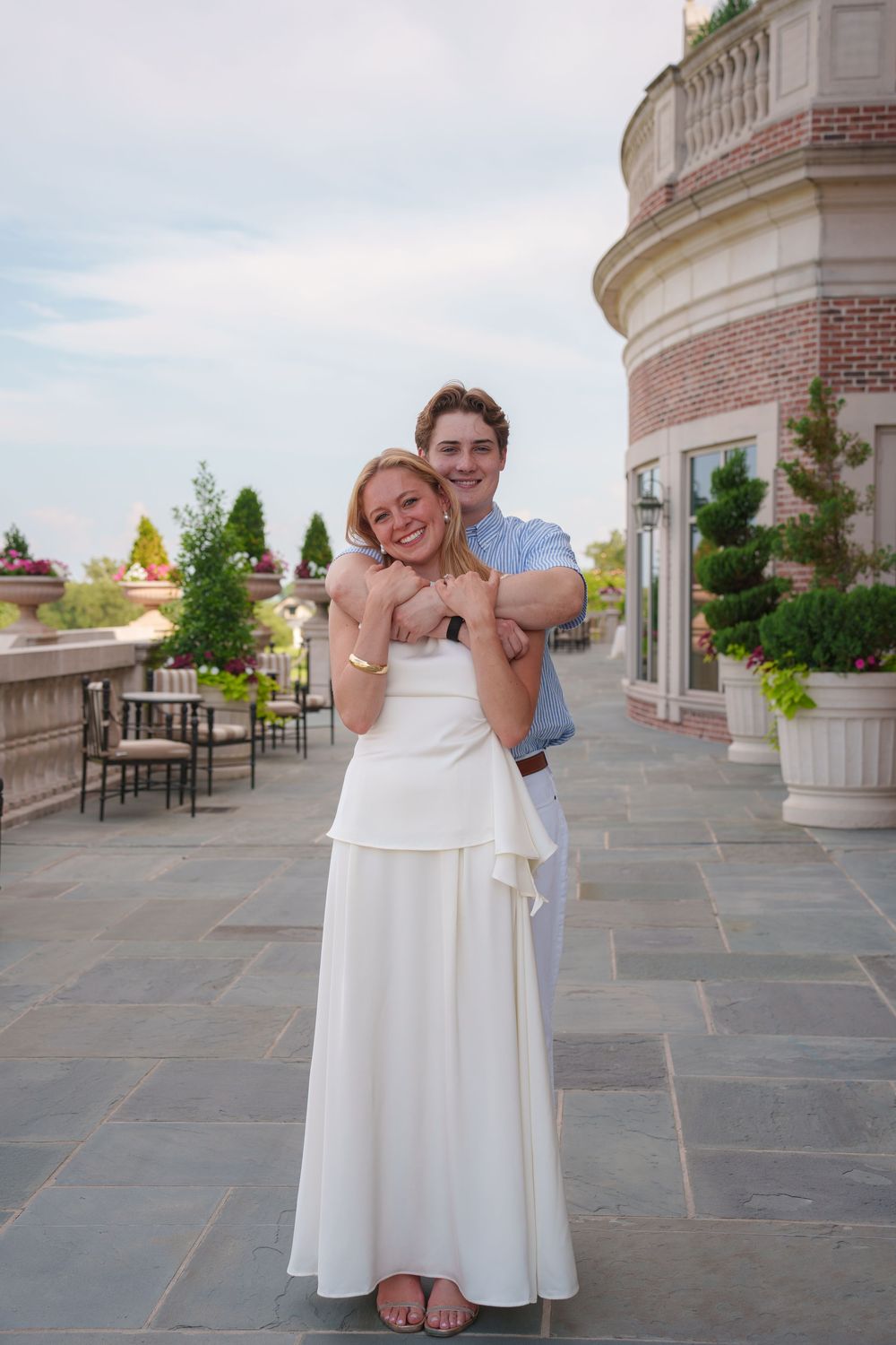 Couple embracing on elegant stone terrace, woman in white gown, man in blue shirt, posing before brick building.