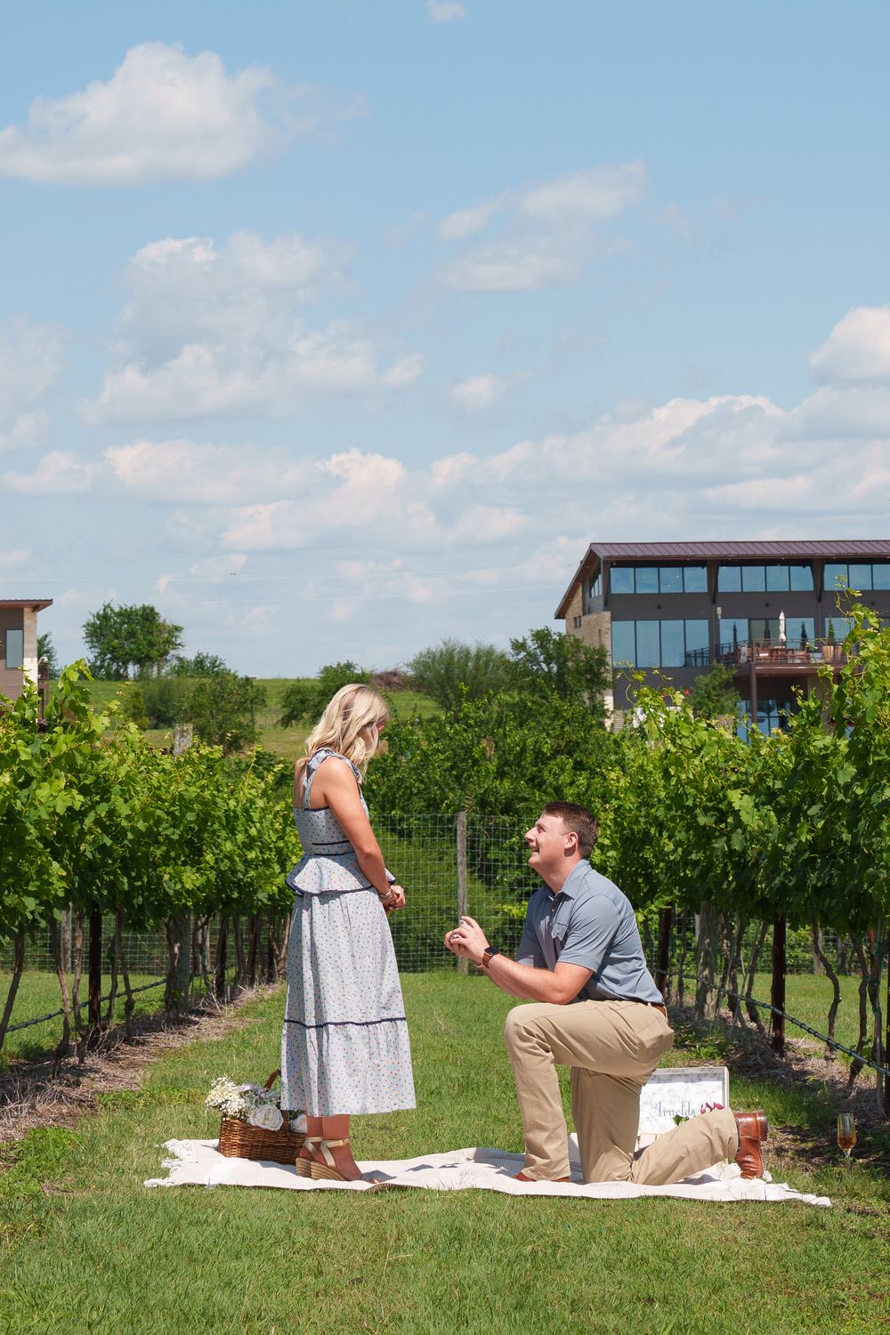 Man proposing on one knee to woman in vineyard during sunny day with winery building in background.