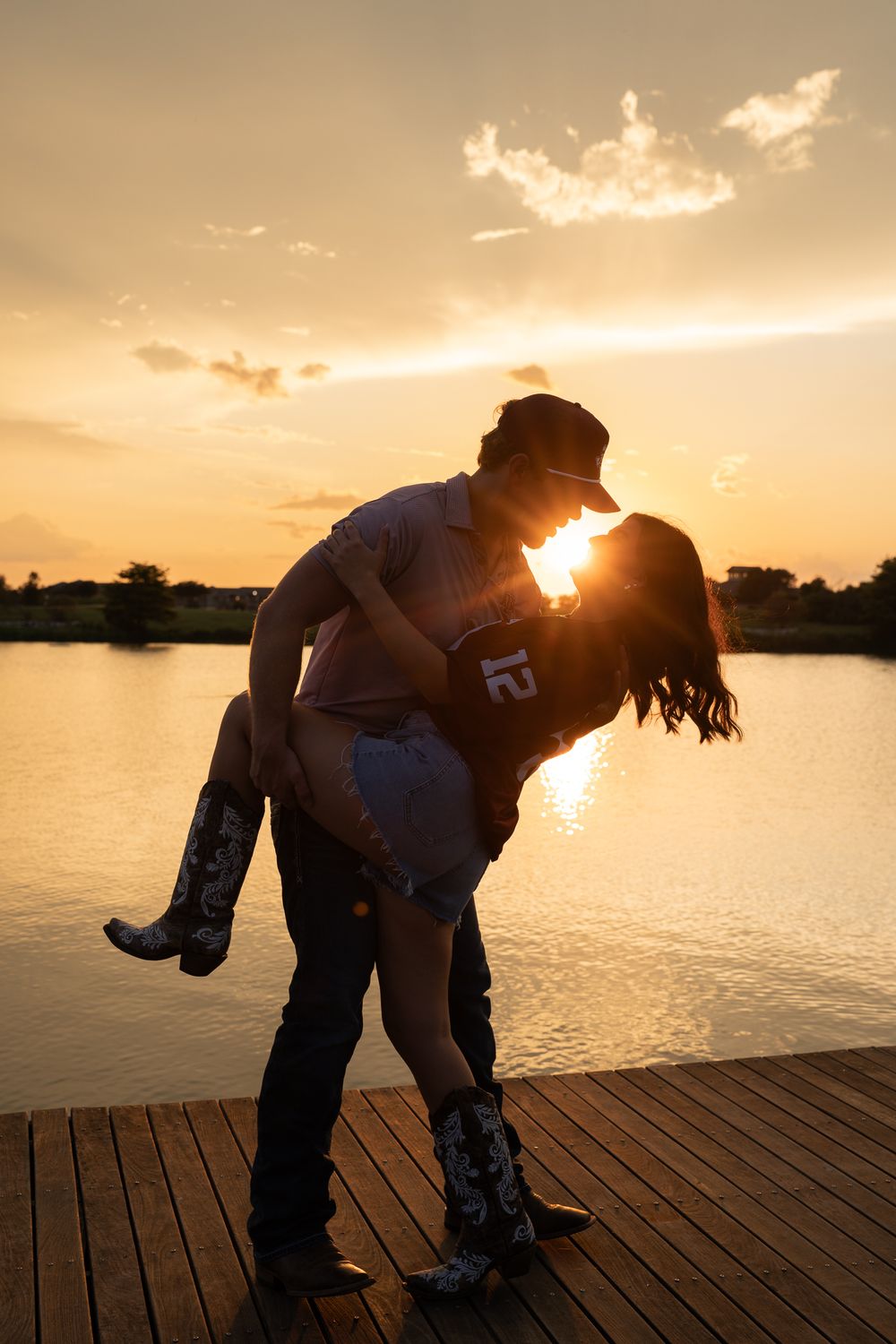 Silhouetted couple shares romantic embrace on lakeside dock during golden sunset while wearing cowboy boots.