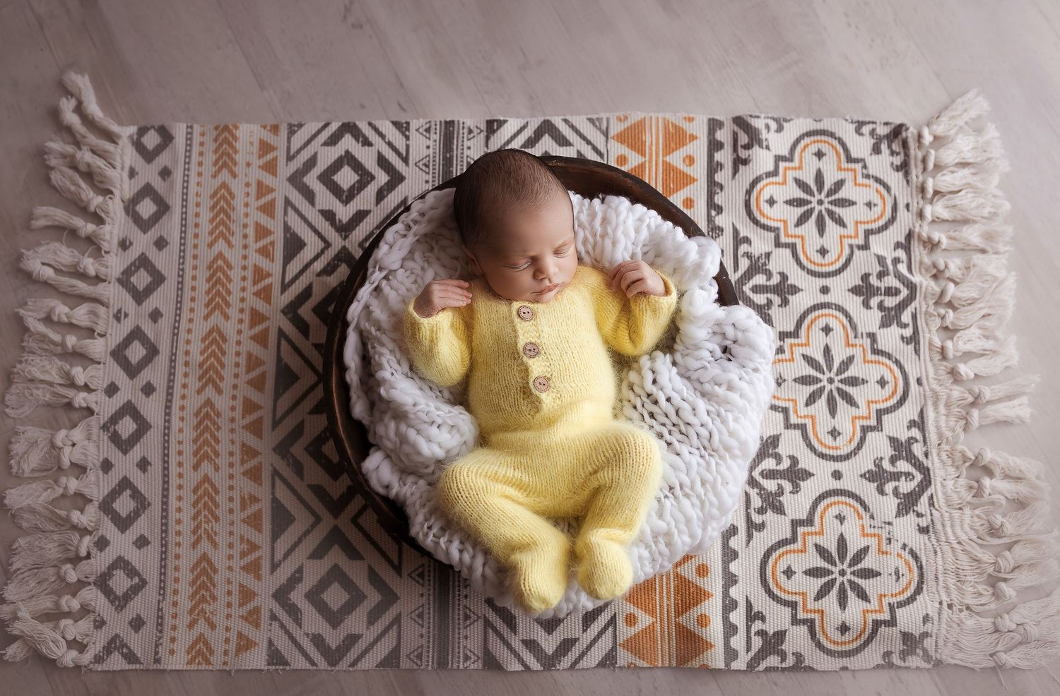 Bebe recien nacido acostado en un bowl de madera con ropita tejida amarilla debajo del bowl hay una alfombra estilo boho Fotografa