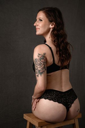 A boudoir portrait showing a woman in black lingerie sitting on a wooden stool with a floral arm tattoo.