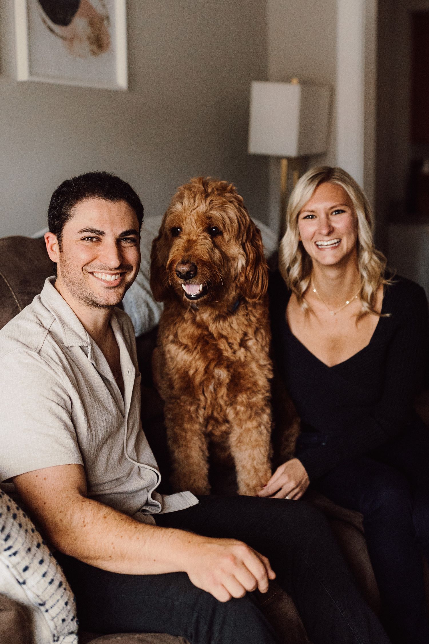 A happy couple poses with their smiling goldendoodle dog in a series of casual indoor photos.