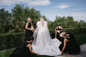 Bridesmaids in black dresses arranging the train of a wedding gown outdoors.